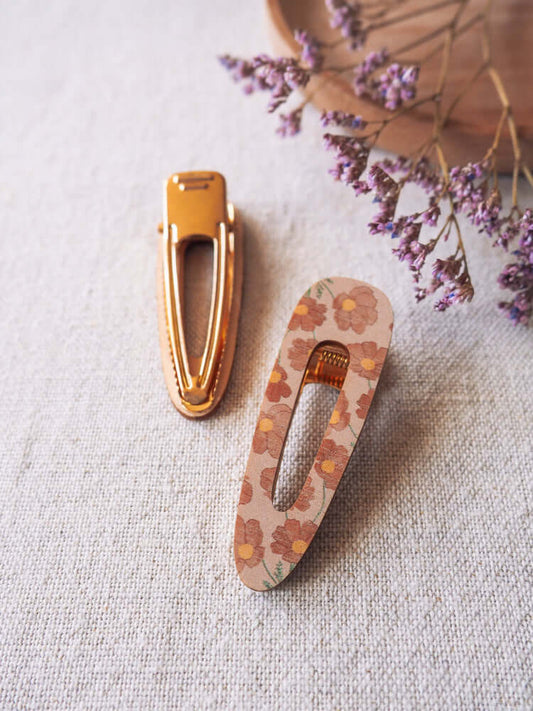 Kaksi vanerista Asta-hiuspinniä tekstiilitaustalla. Pinneissä printti, jossa vaaleanpunaisella taustalla kosmoskukkia. Toinen pinneistä ylösalaisin ja kullan värinen klipsi näkyy. Kuvan yläreunassa puinen lautanen ja violetteja kuivakukkia.